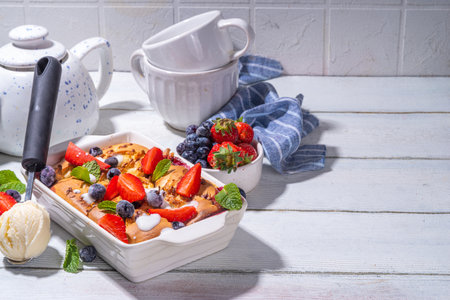 Sweet summer berry cobbler pie, homemade sponge cake with sugared blueberry and strawberry, served with vanilla ice cream ball, on sunny white wooden kitchen tableの写真素材