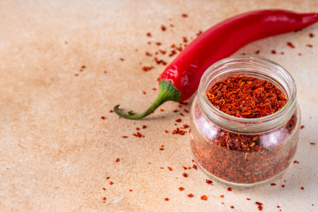 Hot spicy seasoning, powder or flakes red chili pepper in small jar, with fresh chili pepper on beige table. Ingredient for cooking Mexican, Asian spicy dishesの写真素材