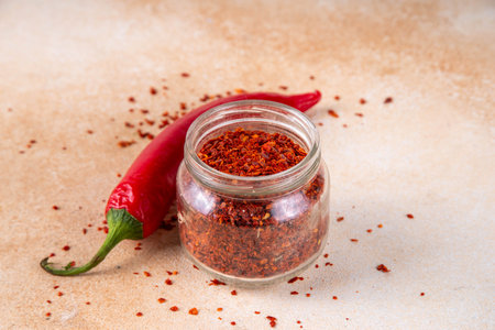 Hot spicy seasoning, powder or flakes red chili pepper in small jar, with fresh chili pepper on beige table. Ingredient for cooking Mexican, Asian spicy dishesの写真素材