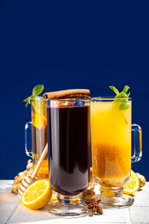 Collection of autumn winter hot drinks. Set of hot lemon ginger tea, mulled wine, fruit citrous punch grog in warm cocktail glass mugs, with cinnamon, spices, dark blue white background copy spaceの写真素材