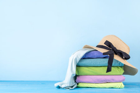 Summer bright clothes pile stack on wooden light blue table, with summer hat, suitcase, shells. Seaside holidays, vacation concept, preparation for travel backgroundの写真素材