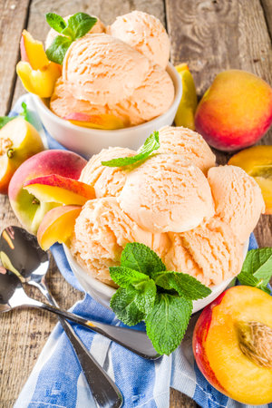 Homemade sweet peach ice cream. Peach gelato balls in small bowls, on wooden background with fresh peaches and mint leavesの写真素材