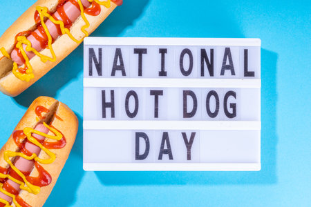 National Hot Dogs day background, hotdog summer party festival foods, Two tasty classic american hot dogs with sauces and lightbox sign with inscription National Hot Dog Day on blue backgroundの写真素材