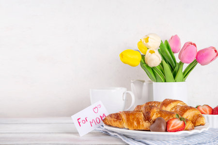 Motherâs Day breakfast background featuring croissant, strawberries, chocolates, coffee mug, bouquet of tulip flowers, as surprise and gift, with greeting card "for mom".の写真素材
