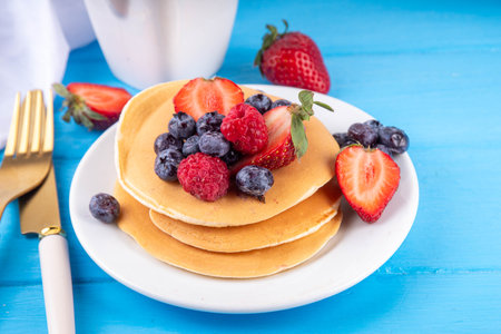 Stack of traditional american pancakes with summer berries. Breakfast sweet pancakes with honey, strawberry and blueberry, on morning sun lighted blue wooden background copy spaceの写真素材