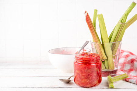 Pink and green rhubarb sweet jam. Homemade tasty sweet rhubarb confiture preserves, in small jar on white wooden table, with fresh rhubarb in the backgroundの写真素材