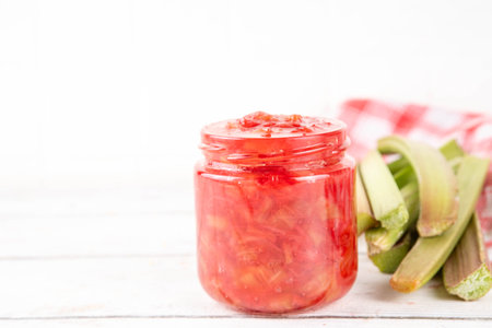 Pink and green rhubarb sweet jam. Homemade tasty sweet rhubarb confiture preserves, in small jar on white wooden table, with fresh rhubarb in the backgroundの写真素材