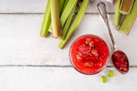 Pink and green rhubarb sweet jam. Homemade tasty sweet rhubarb confiture preserves, in small jar on white wooden table, with fresh rhubarb in the backgroundの写真素材
