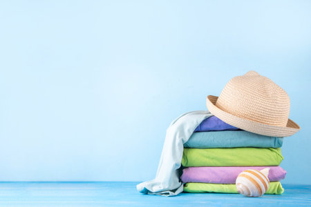 Summer bright clothes pile stack on wooden light blue table, with summer hat, suitcase, shells. Seaside holidays, vacation concept, preparation for travel backgroundの写真素材