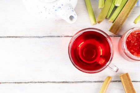 Organic natural rhubarb tea, sweet herbal plant based pink and green rhubarb hot tea drink on teapot and glass cup, on white wooden table with fresh rhubarb copy spaceの写真素材