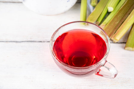 Organic natural rhubarb tea, sweet herbal plant based pink and green rhubarb hot tea drink on teapot and glass cup, on white wooden table with fresh rhubarb copy spaceの写真素材