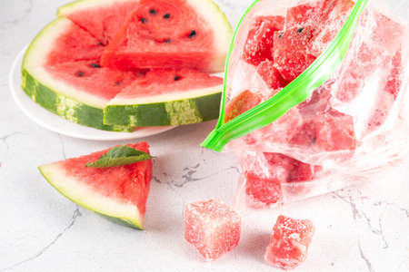 Frozen watermelon slices, cubes in freezing bags with fresh ripe raw watermelon on kitchen white table, Healthy simple canning, freezing stock up of fruit harvest for winter concept.の写真素材