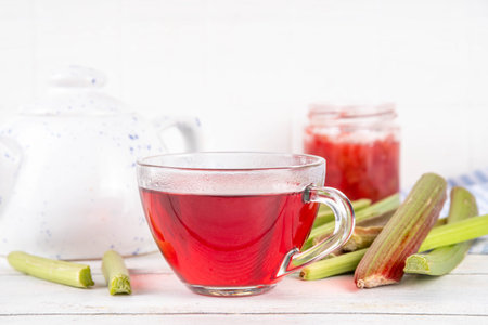 Organic natural rhubarb tea, sweet herbal plant based pink and green rhubarb hot tea drink on teapot and glass cup, on white wooden table with fresh rhubarb copy spaceの写真素材