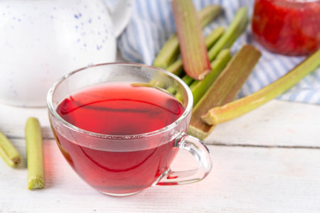 Organic natural rhubarb tea, sweet herbal plant based pink and green rhubarb hot tea drink on teapot and glass cup, on white wooden table with fresh rhubarb copy spaceの写真素材