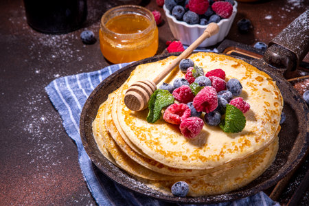 Homemade french tiny pancakes, battercakes with berry. Stack of freshly baked thin crepes on frying pan with fresh blueberry, raspberries and honey. Healthy morning breakfast on dark backgroundの写真素材