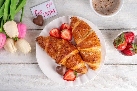 Motherâs Day breakfast background featuring croissant, strawberries, chocolates, coffee mug, bouquet of tulip flowers, as surprise and gift, with greeting card "for mom".の写真素材