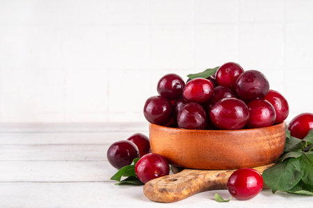 Fresh raw ripe cherry plum with leaves. Bowl of tasty farm bright red cherry plum fruits, in wooden craft bowl on white wooden background copy space