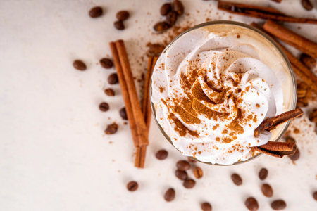 Iced cinnamon latte with whipped cream, cinnamon syrup and cinnamon powder on top, with coffee beans on white and beige background copy spaceの写真素材