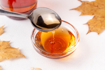 Tasty golden maple syrup in bottle, bowl and spoon, with maple leaves on white background, copy spaceの写真素材