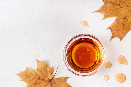 Tasty golden maple syrup in bottle, bowl and spoon, with maple leaves on white background, copy spaceの写真素材