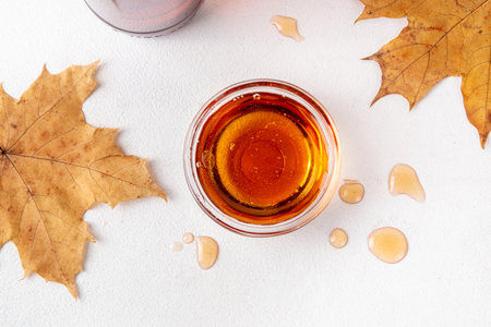 Tasty golden maple syrup in bottle, bowl and spoon, with maple leaves on white background, copy spaceの写真素材