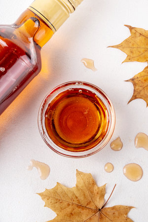Tasty golden maple syrup in bottle, bowl and spoon, with maple leaves on white background, copy spaceの写真素材