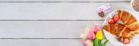 Motherâs Day breakfast background featuring croissant, strawberries, chocolates, coffee mug, bouquet of tulip flowers, as surprise and gift, with greeting card "for mom".の写真素材