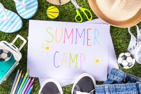 Kids summer camp invitation background. Bright yellow backdrop with Summer Camp ad text banner, pencils, summer shoes, flip-flops, ball, sneakers, sand toys, summer hat, suitcase, on grass background
