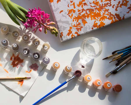 on a white table the brushes and canvas are laid out for drawing on numbers nearby there is a geocint flower and a glass with water that casts a beautiful shadowの写真素材