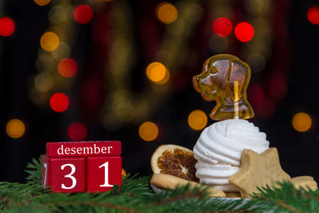 red cubes calendar date and date December 31, tangerines and a plate of sweets with marshmallow and caramel as a dog, dried mandarin, gingerbread on fir branches against a background of yellow and red bokeh lights on a black backgroundの写真素材