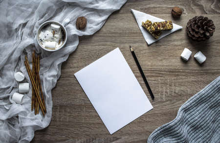 on a wooden background a white sheet and a hand write in black pencil on paper, and on the left a marshmallow and sweet straw strips, cloth, walnuts, a cone, a muesli bar on a white triangular napkin, a mug with cocoa, in which lies a melted snowman from marshmallowsの写真素材