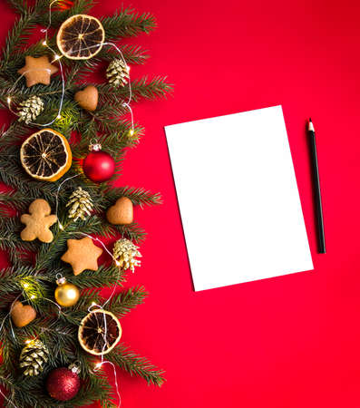 on a red background green spruce branches decorated with gingerbreads, golden cones, oranges, lights and balls and on the side is a white sheet and a black pencilの写真素材