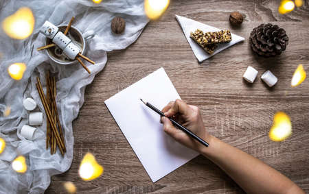 on a wooden background a white sheet and a hand write in black pencil on paper, and on the left a marshmallow and sweet straw stripe, chopsticks, cloth, walnuts, a cone, a muesli bar on a white triangular napkin, the lights of a garland around, a mug with cocoa, in which lies a snowman from marshmallowsの写真素材