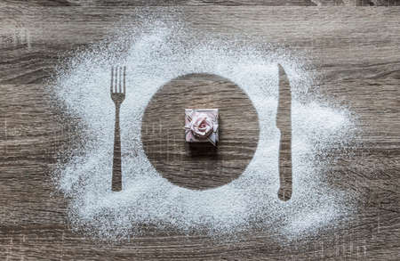 on a wooden background with powdered powder snow covered with silhouette a plate a fork knife of appliances in the center lies a pink gift box with a bowの写真素材