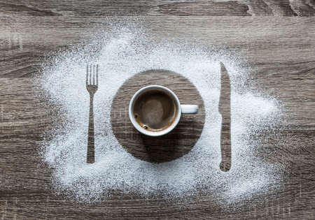 on a wooden background with powdered powder snow covered with a silhouette a plate fork knife of appliances in the center is a white cup with hot coffee with foamの写真素材