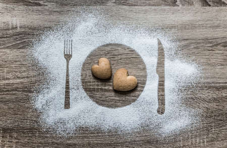 on a wooden background with powdered powder snow covered with a silhouette a plate a fork knife of appliances in the center lies two cookies in the form of a heartの写真素材