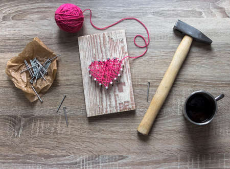 on a wooden table lies a hammer of a nail of a thread pink and a gift with your hands a heart of threads and nails a cup of coffeeの写真素材