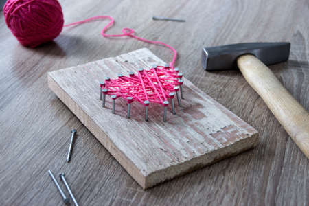 on a wooden table lies a hammer of a nail of a thread pink and a gift with your hands a heart of threads and nailsの写真素材