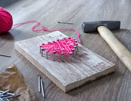 on a wooden table lies a hammer of a nail of a thread pink and a gift with your hands a heart of threads and nailsの写真素材