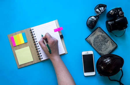 on blue blue background hand writing pen in notebook on springs notes stickers colored side glasses bag handbag coco phone passport map cameraの写真素材