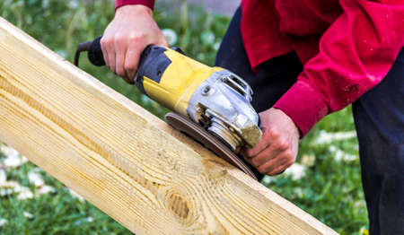 male hands in a dark red shirt hold a yellow grinder and process a wooden boardの写真素材