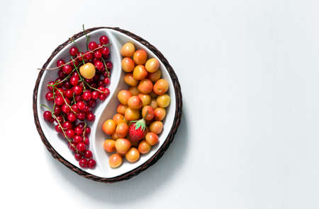 on white background round bowl of yin yang with summer berries of red currant and yellow cherry green leaf of strawberryの写真素材