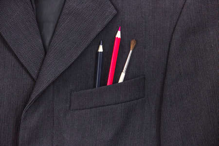 in the breast pocket of a dark black man's jacket a pencil and a brush lay in a light strip red blueの写真素材