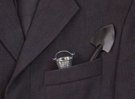 In a breast pocket of a dark black man's jacket in a light strip there is a bucket and a shovelの写真素材