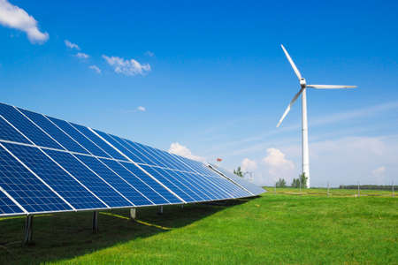 Wind generator of electricity from three blades and solar panels of a battery of photocells against a background of clouds and a blue sky green grassの写真素材