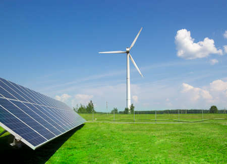 wind generator of electricity from three blades and solar panels of a battery of photocells against a background of clouds and a blue sky green grassの写真素材