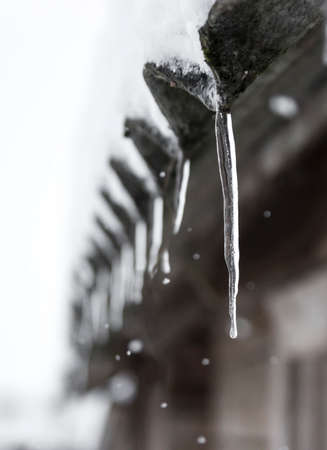 icicles hang and snow falls on the roof of a wooden houseの写真素材