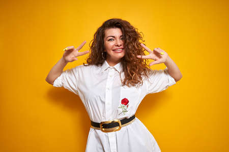 Don't worry, be happy. A natural portrait of a young girl with curly hair on a yellow background. She smiles and holds her hands in a world peace style.の写真素材