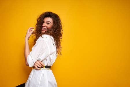 Portrait of a young beautiful girl with curly lush hair in a white shirt on a yellow background. She smiles and stands sideways. It stands on the left side of the frame. Laughs emotionally.の写真素材