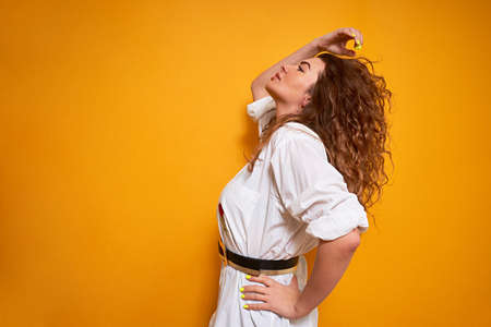 A young curly-haired girl with a full head of hair is standing sideways on a yellow background and looking up. With one hand on his belt and the other straightening his hair.の写真素材